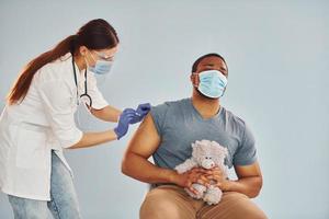 impaurito tipo con orsacchiotto orso. femmina medico dando iniezione per africano americano uomo a ospedale foto