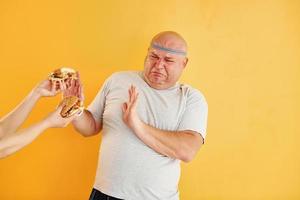 mani con hamburger tenta tipo. divertente sovrappeso uomo nel allegro testa cravatta è contro giallo sfondo foto