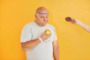 Mela o biscotto. divertente sovrappeso uomo nel allegro testa cravatta è contro giallo sfondo foto