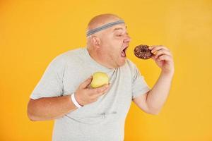 Mela o biscotto. divertente sovrappeso uomo nel allegro testa cravatta è contro giallo sfondo foto