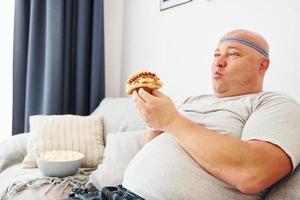 si siede su il divano con Popcorn e Hamburger. divertente sovrappeso uomo nel casuale Abiti è in casa a casa foto