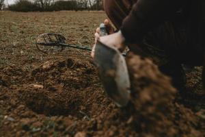 dettaglio di dell'uomo mani con pala nel esso, durante scavando un' buco nel terra dopo positivo segnale di metallo rivelatore con dire bugie metallo rivelatore nel sfondo foto