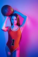 nel pallacanestro uniforme. alla moda giovane donna in piedi nel il studio con neon leggero foto