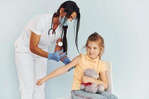 poco ragazza con orsacchiotto orso. medico nel uniforme fabbricazione vaccinazione per il paziente foto