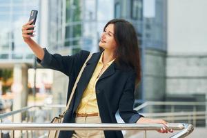 Tenere Telefono nel mano. donna nel formale indossare in piedi all'aperto nel il città a giorno foto