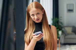 in piedi con Telefono. femmina adolescente con bionda capelli è a casa a giorno foto