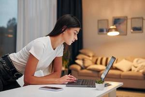 utilizzando il computer portatile. donna nel bianca camicia e nero jeans è in piedi in casa a casa foto