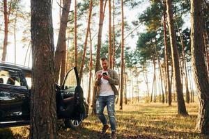 vicino il macchina. uomo nel jeans è all'aperto nel il foresta con il suo nero colorato automobile foto