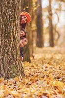 nascondiglio dietro a albero. madre con sua poco figlio e figlia è avendo divertimento nel il autunno parco foto