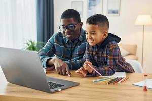 con il computer portatile su tavolo. africano americano padre con il suo giovane figlio a casa foto