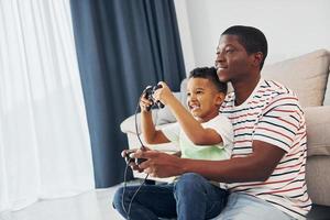 giocando video Giochi. africano americano padre con il suo giovane figlio a casa foto