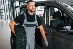 con pneumatico nel mani. uomo nel uniforme è Lavorando nel il autosalone a giorno foto