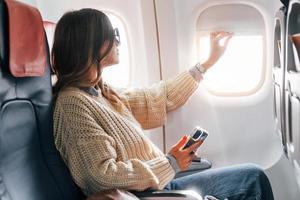 seduta con Telefono nel mani. giovane femmina passanger nel casuale Abiti è nel il aereo foto
