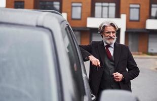 alla moda anziano uomo con grigio capelli e barba in piedi all'aperto su il strada vicino il suo auto con chiavi nel mano foto