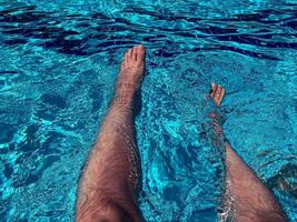 rilassante a il nuoto piscina. uomo rilassante. uomo godendo il caldo estate a piscina. prendere il sole di il nuoto piscina, mans gambe dire bugie giù su un' sole lettino al di sopra di il acqua foto