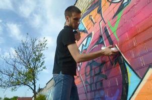 giovane graffiti artista con zaino e gas maschera su il suo collo vernici colorato graffiti nel rosa toni su mattone parete. strada arte e contemporaneo pittura processi foto