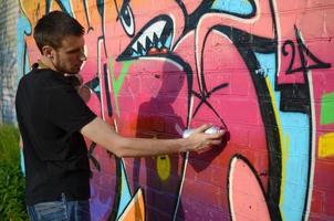 giovane graffiti artista con zaino e gas maschera su il suo collo vernici colorato graffiti nel rosa toni su mattone parete. strada arte e contemporaneo pittura processi foto