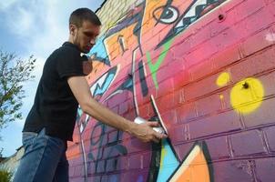 giovane graffiti artista con zaino e gas maschera su il suo collo vernici colorato graffiti nel rosa toni su mattone parete. strada arte e contemporaneo pittura processi foto