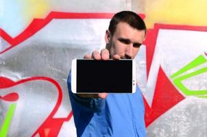 il graffiti artista dimostra un' smartphone con un vuoto nero schermo contro il sfondo di un' colorato dipinto parete. strada arte concetto foto