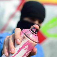 un' giovane graffiti artista nel un' blu giacca e nero maschera è Tenere un' può di dipingere nel davanti di lui contro un' sfondo di colorato graffiti disegno. strada arte e vandalismo concetto foto