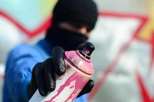 un' giovane graffiti artista nel un' blu giacca e nero maschera è Tenere un' può di dipingere nel davanti di lui contro un' sfondo di colorato graffiti disegno. strada arte e vandalismo concetto foto