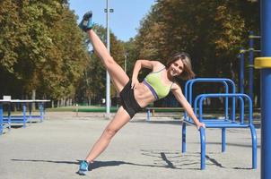 un' giovane caucasico femmina atleta nel luminosa verde sport Abiti praticante gamba cavalcate su il gli sport Palestra per attivo all'aperto ricreazione. estate gli sport e salutare stile di vita foto