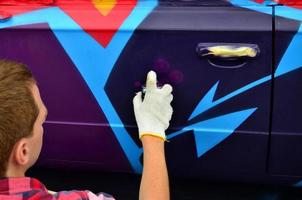 un' giovane dai capelli rossi graffiti artista vernici un' nuovo colorato graffiti su il macchina. foto di il processi di disegno un' graffiti su un' auto avvicinamento. il concetto di strada arte e illegale vandalismo