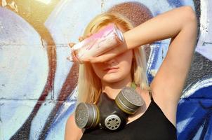 giovane e bellissimo sexy ragazza graffiti artista con gas maschera su sua collo nascondiglio il suo occhi con un' spray può in piedi su un' parete sfondo con un' graffiti modello nel blu e viola toni foto