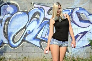 un' giovane e bellissimo sexy ragazza graffiti artista con un' dipingere spray e gas maschera su sua collo sta su il parete sfondo con un' graffiti modello nel blu e viola toni foto