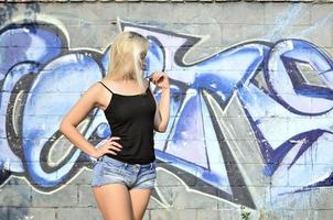 sexy caucasico bionda ragazza nel denim pantaloncini e nero serbatoio superiore in posa contro graffiti parete nel il giorno all'aperto foto