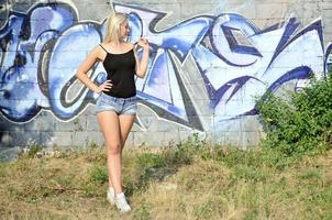 sexy caucasico bionda ragazza nel denim pantaloncini e nero serbatoio superiore in posa contro graffiti parete nel il giorno all'aperto foto