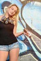 un' giovane e bellissimo sexy ragazza graffiti artista con un' dipingere spray e gas maschera su sua collo sta su il parete sfondo con un' graffiti modello nel blu e viola toni foto