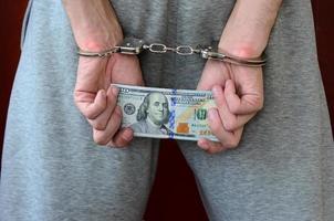 un arrestato tipo nel grigio pantaloni con Ammanettato mani detiene un' enorme quantità di dollaro fatture. indietro Visualizza foto