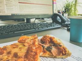 ufficio lavoratore merenda. vicino il Lavorando computer e altro ufficio attrezzatura bugie un' delizioso pezzo di Pizza con Riempimento. Pizza su Impasto con carne e formaggio foto