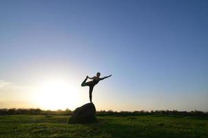 silhouette di giovane bionda ragazza nel sport completo da uomo pratica yoga su un' pittoresco verde collina nel il sera a tramonto. il concetto di esercizio e salutare stili di vita foto