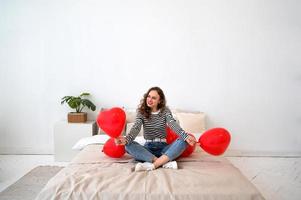 San Valentino giorno. giovane contento femmina su il letto con rosso cuore sagomato palloncini foto