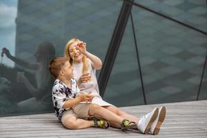 contento biondo donna e poco ragazzo seduta su terrazza e mangiare dolci. madre e figlio godere tempo insieme. positivo giovane mamma giocando, la spesa tempo con sua carino bambino, ridendo, avendo divertimento. famiglia. foto