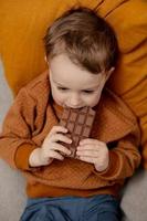 poco adorabile ragazzo seduta su il divano a casa e mangiare cioccolato sbarra. bambino e dolci, zucchero confetteria. ragazzo godere un' delizioso dolce. prescolastico bambino con casuale vestiario. positivo emozione. foto