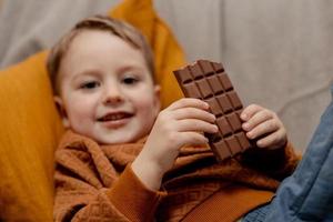 poco adorabile ragazzo seduta su il divano a casa e mangiare cioccolato sbarra. bambino e dolci, zucchero confetteria. ragazzo godere un' delizioso dolce. prescolastico bambino con casuale vestiario. positivo emozione. foto