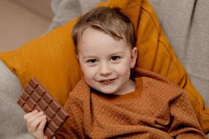 poco adorabile ragazzo seduta su il divano a casa e mangiare cioccolato sbarra. bambino e dolci, zucchero confetteria. ragazzo godere un' delizioso dolce. prescolastico bambino con casuale vestiario. positivo emozione. foto