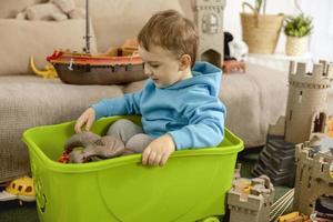 poco caucasico ragazzo con blu felpa con cappuccio giocando con colorato giocattoli a casa. bambino avendo divertimento. contento e allegro ragazzo giochi con nave, dinosauri, castello. tempo libero attività, domestico vita. accogliente camera. foto