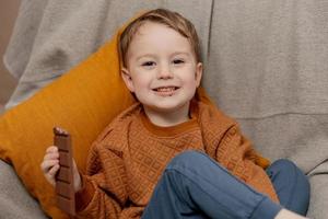 poco adorabile ragazzo seduta su il divano a casa e mangiare cioccolato sbarra. bambino e dolci, zucchero confetteria. ragazzo godere un' delizioso dolce. prescolastico bambino con casuale vestiario. positivo emozione. foto