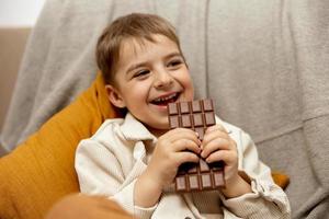 poco adorabile ragazzo seduta su il divano a casa e mangiare cioccolato sbarra. bambino e dolci, zucchero confetteria. ragazzo godere un' delizioso dolce. prescolastico bambino con casuale vestiario. positivo emozione. foto