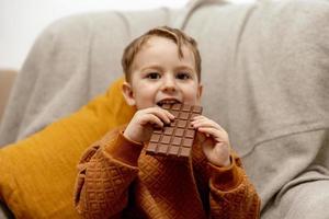 poco adorabile ragazzo seduta su il divano a casa e mangiare cioccolato sbarra. bambino e dolci, zucchero confetteria. ragazzo godere un' delizioso dolce. prescolastico bambino con casuale vestiario. positivo emozione. foto