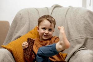 poco adorabile ragazzo seduta su il divano a casa e mangiare cioccolato sbarra. bambino e dolci, zucchero confetteria. ragazzo godere un' delizioso dolce. prescolastico bambino con casuale vestiario. positivo emozione. foto