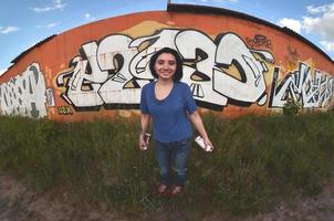 ritratto di un emotivo giovane ragazza con nero capelli e piercing. un' angolo ampio foto di un' ragazza con aerosol dipingere lattine nel il mani su un' graffiti parete sfondo. un' moderno ritratto di un' fisheye lente