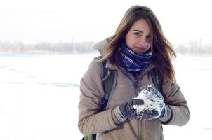 un' giovane e gioioso caucasico ragazza nel un' Marrone cappotto detiene un' palla di neve nel il sfondo di un' orizzonte linea fra il cielo e un' congelato lago nel inverno foto