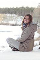 un' giovane caucasico ragazza nel un' Marrone cappotto è seduta vicino un' scogliera nel il sfondo di un' orizzonte linea fra il cielo e un' congelato lago nel inverno tempo foto