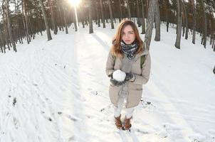 un' giovane e gioioso caucasico ragazza nel un' Marrone cappotto detiene un' palla di neve nel un' innevato foresta nel inverno. fisheye foto