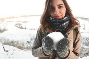 un' giovane e gioioso caucasico ragazza nel un' Marrone cappotto detiene un' palla di neve nel davanti di un' orizzonte linea fra il cielo e un' congelato lago nel inverno. fisheye foto